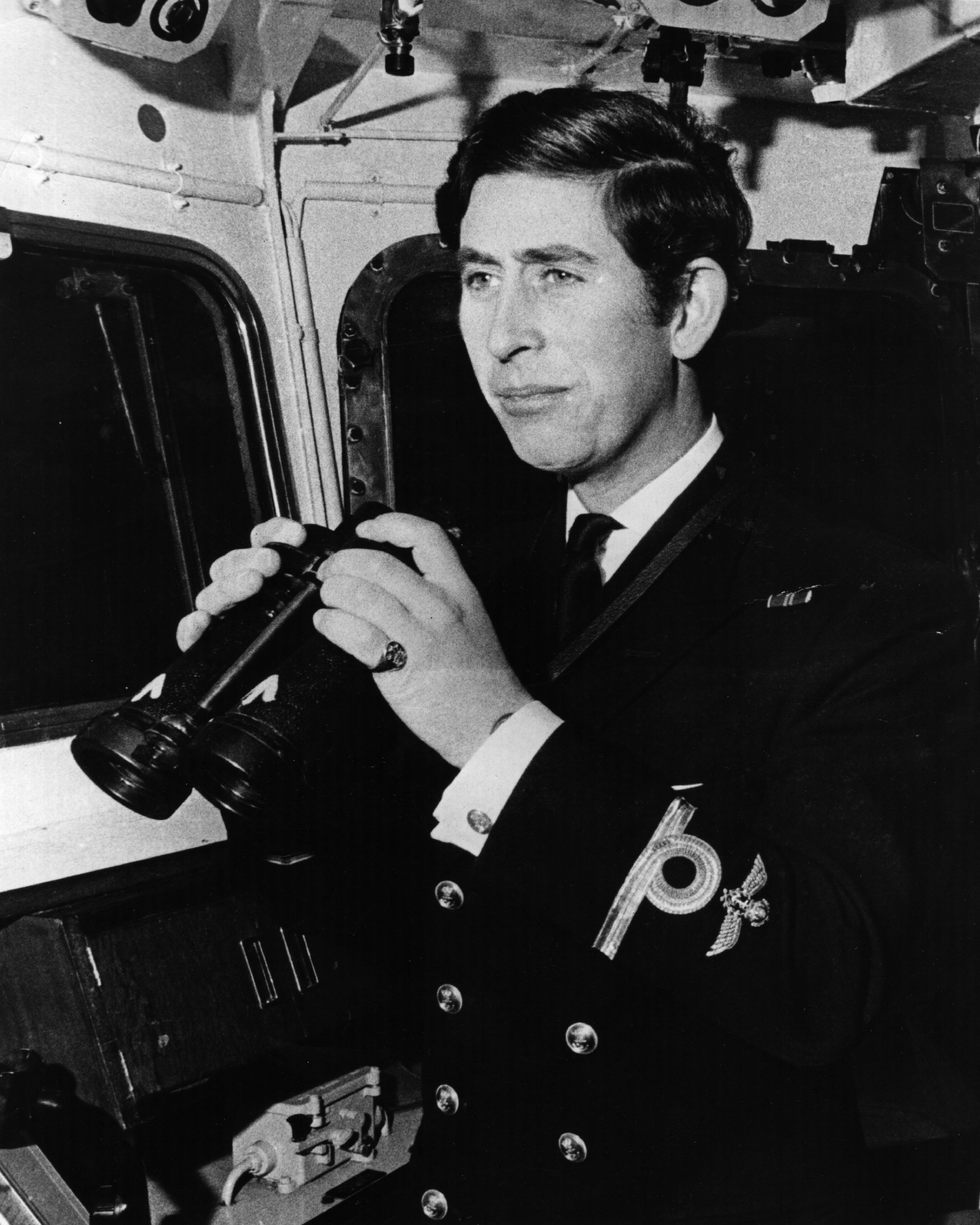 A young Prince Charles (now King Charles III) peers through binoculars aboard a Royal Navy vessel during his service in the 1970s.